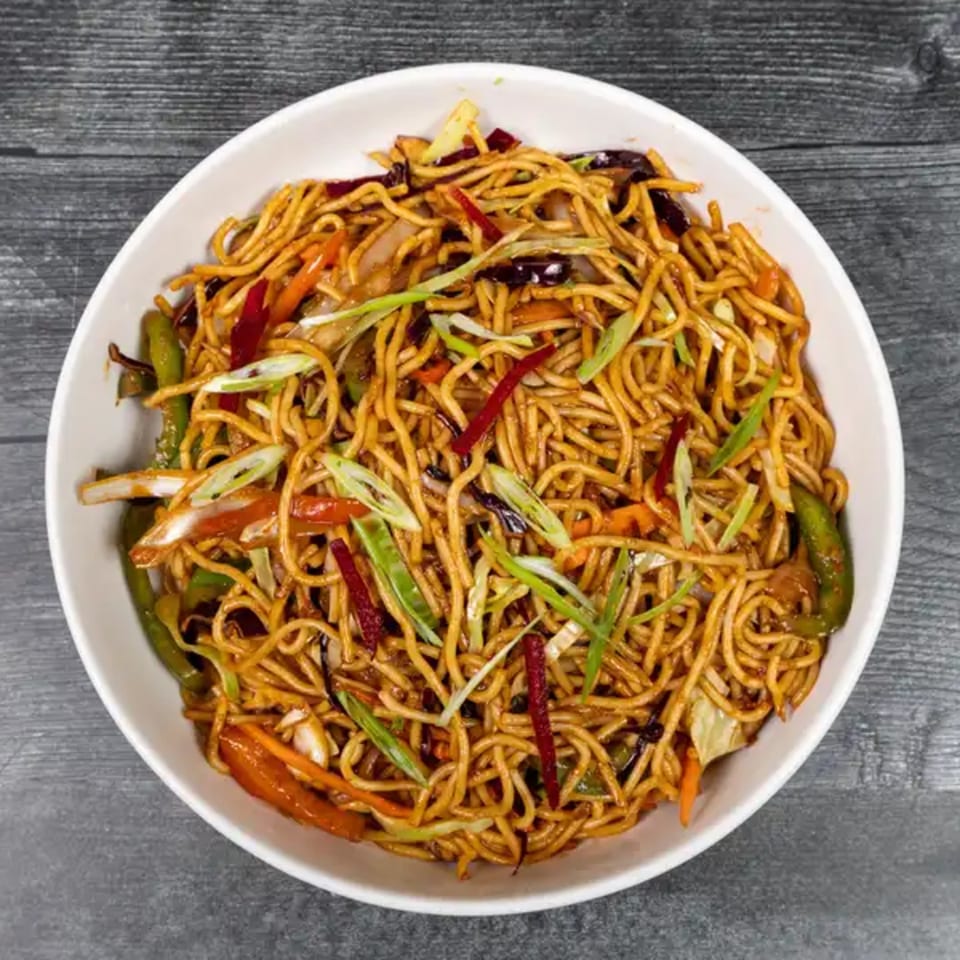 A white bowl filled with Indian Hakka noodles, garnished with colorful vegetables and spring onions.