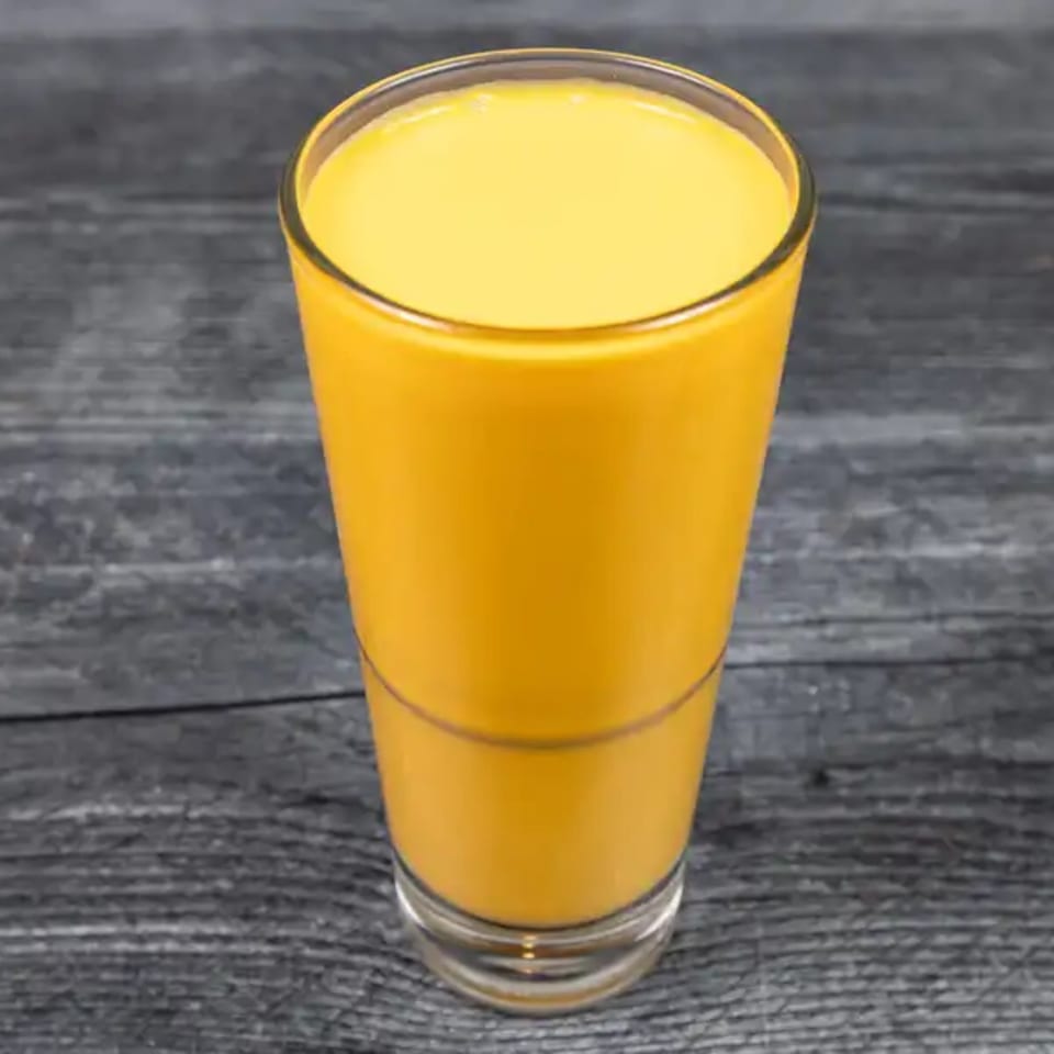 A tall glass of bright yellow mango lassi, an Indian yogurt drink, on a dark wooden surface.
