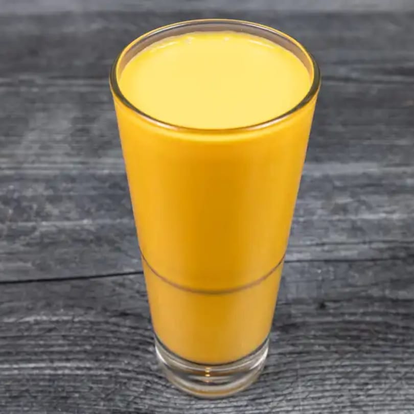 A tall glass of bright yellow mango lassi, an Indian yogurt drink, on a dark wooden surface.