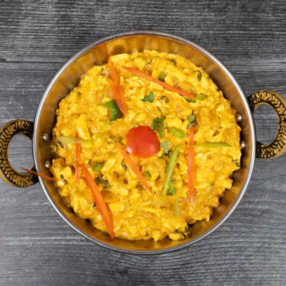 Indian scrambled paneer dish, garnished with red and green peppers and a cherry tomato, in a metal bowl.