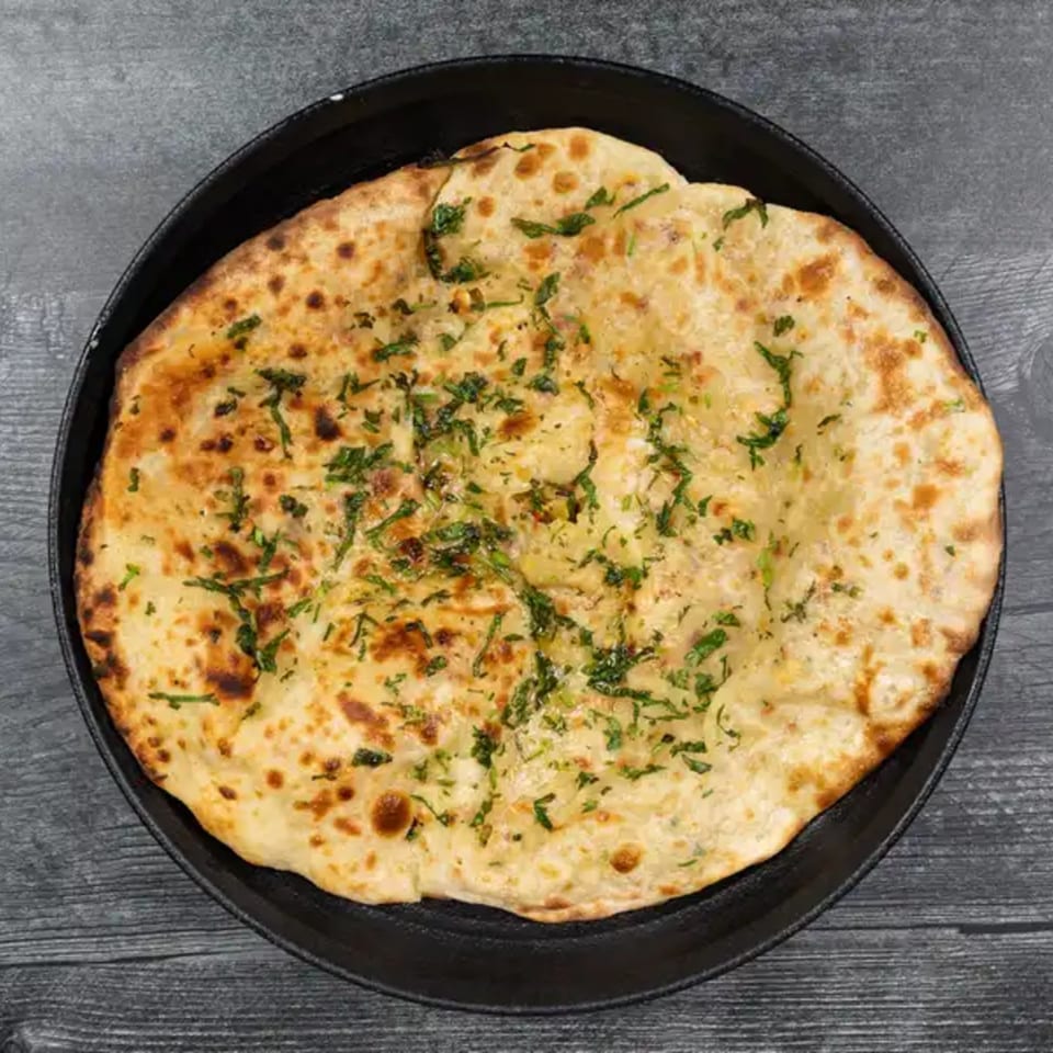 Golden-brown garlic naan bread, topped with fresh green herbs, served in a black pan.